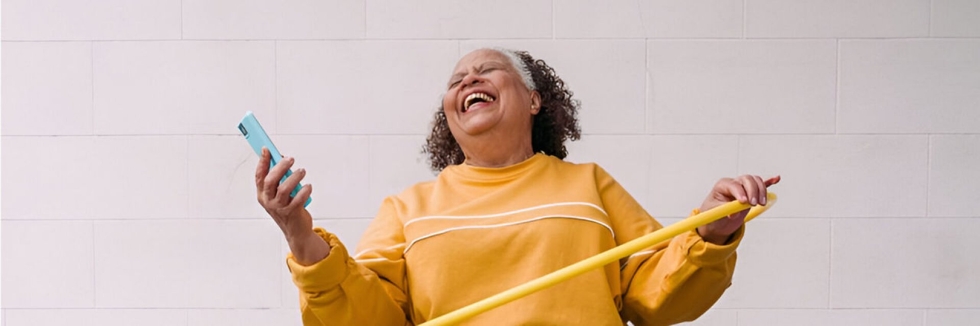 A woman in a yellow sweater laughs joyfully, holding a smartphone and a yellow hula hoop against a plain wall.