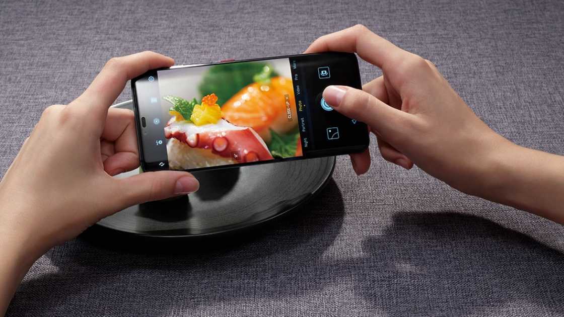A person taking a photograph of sushi on a black plate with a Huawei Mate 20 Pro, against a grey background.