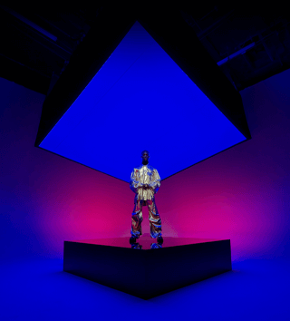 A person dressed in a metallic suit standing on a square centre stage and encompassing the background showcasing the Ultra Wide camera on iPhone 16 Pro Max