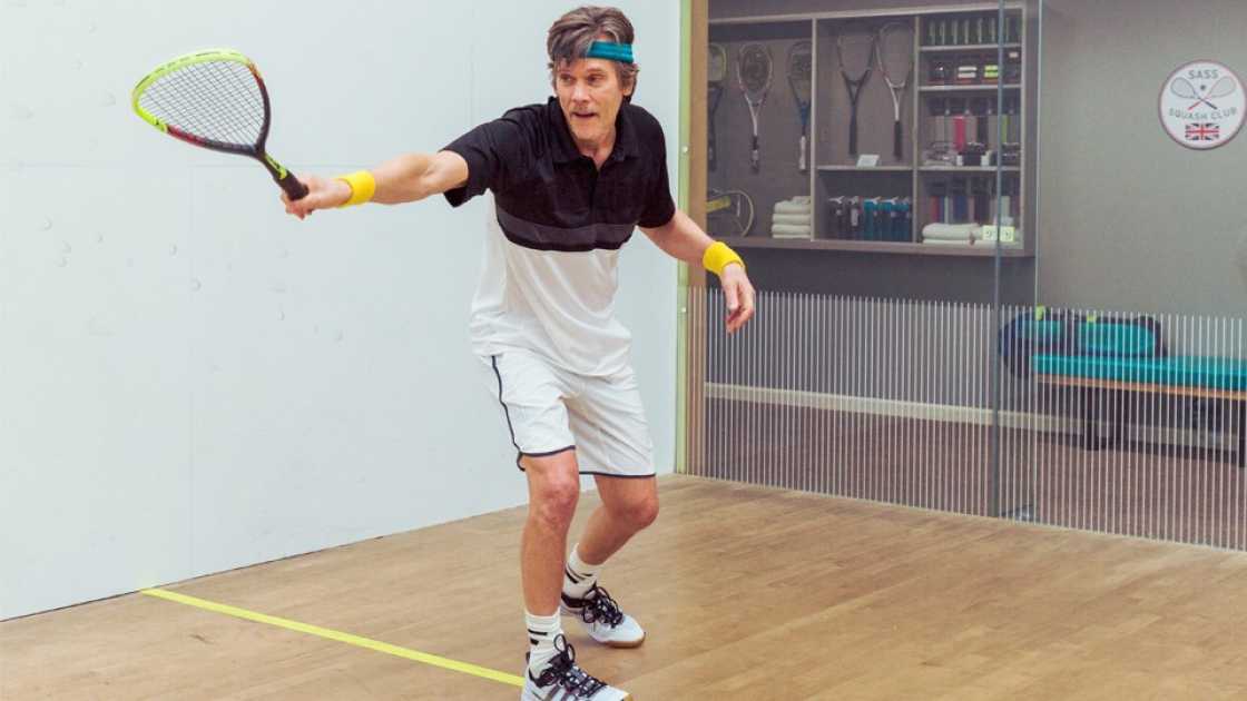 Kevin Bacon extending his arm whilst holding a squash racket 