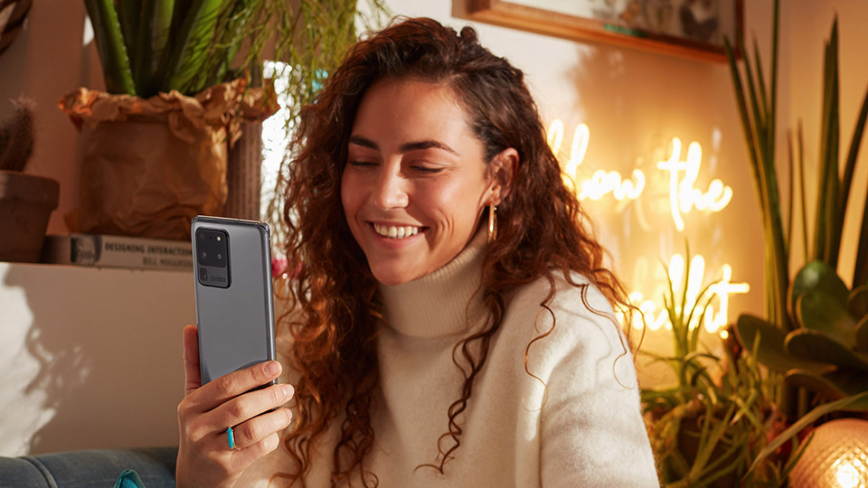 a woman with curly brown hair looking at her Galaxy S20 Ultra 5G while smiling and sitting in a cosy indoor setting
