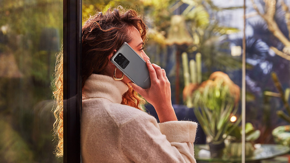 a woman with brown curly hair stood by the window talking on her samsung galaxy s20 ultra 5g while looking outdoors