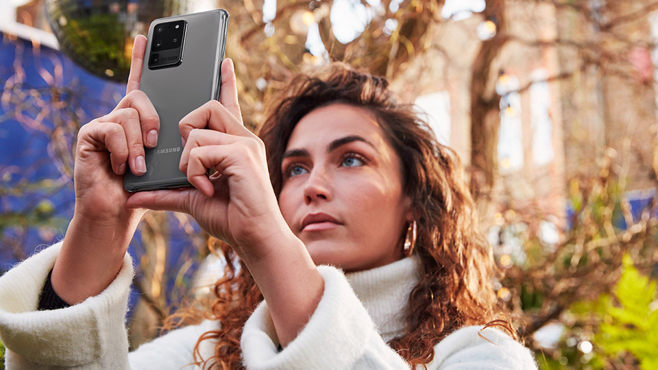 a woman with brown curly hair holding up her Samsung Galaxy S20 Ultra 5G to take a photo in an outdoor setting, with a disco ball in the background