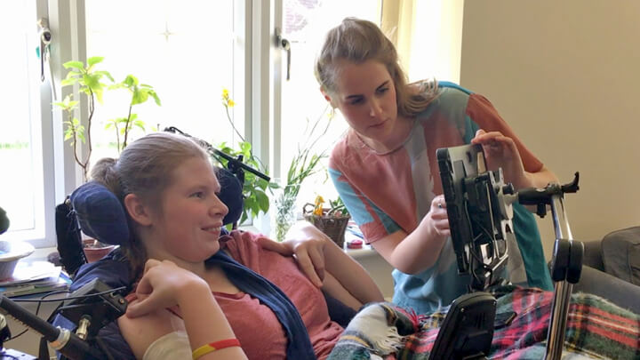 A physically disabled woman is playing video games using customised equipment. Another woman is showing her something on the monitor