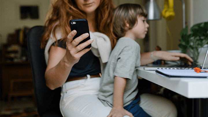 A woman studies her phone while working, with her young son sat on her lap